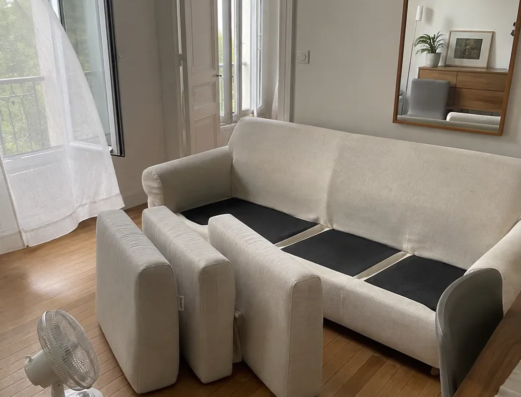 Canapé en tissu écru dont les coussins sèchent debout, fenêtre ouverte et petit ventilateur au sol