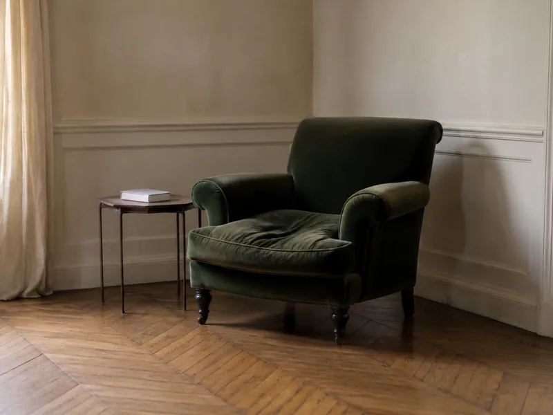 Fauteuil en velours vert foncé dans un angle de salon, à côté d'une petite table et d'un rideau en lin